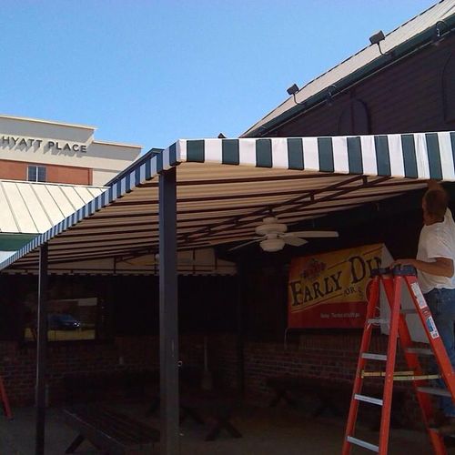 A man finishes construction on a small patio cover outside of a building. Several benches are scattered about the patio. A building in the distance reads 'Hyatt Place'