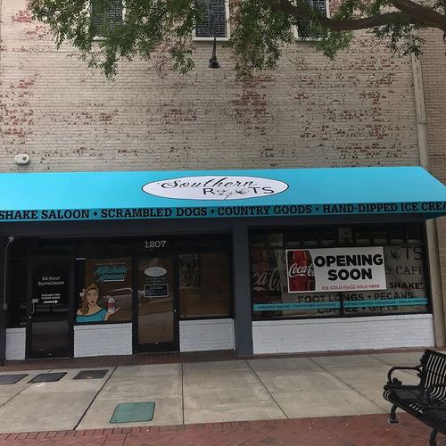 A large awning covering a storefront reads 'Southern Roots - Shade Saloon - Scrambled Dogs - Country Goods - Hand-dipped Ice Cream'. A sign on the window indicates the store will be opening soon.