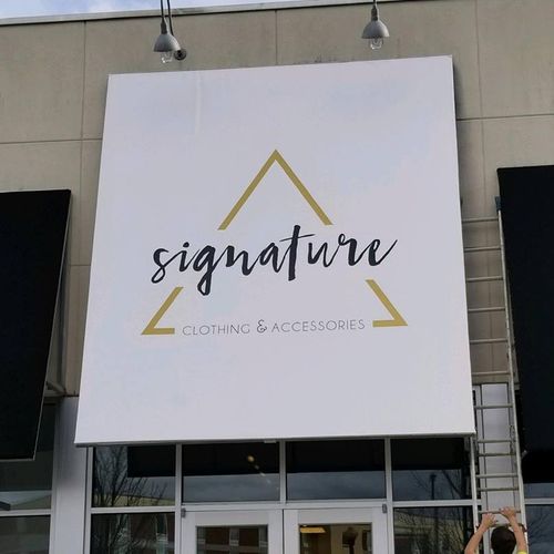 A tall awning covers the entrance to the 'Signature - Clothing & Accessories' building. An installer on a ladder is just finishing work on it.