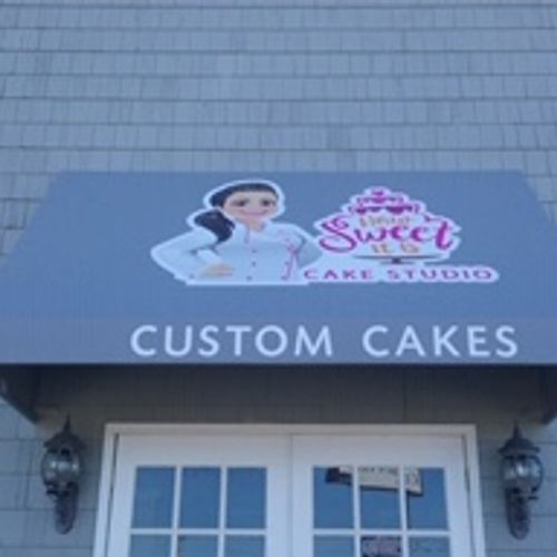 An awning over the door to a business with an image of a female baker reads 'How Sweet Cake Studio - Custom Cakes'